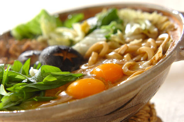 ベージュ色の土鍋に入った、しいたけ、卵、野菜などの味噌煮込みうどん