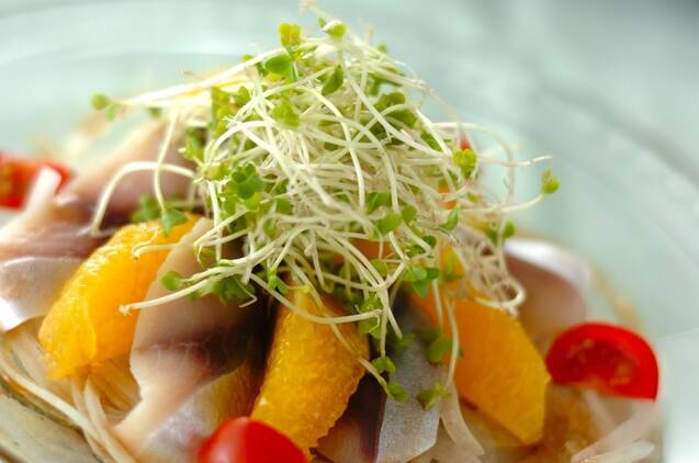 透明のガラス皿にしめサバとオレンジのサラダが盛られ、机の上に置かれている