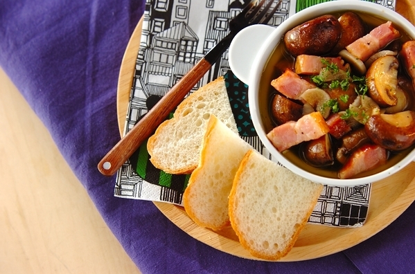 木のプレートにのっているスープカップに入ったアヒージョとバゲットとフォーク