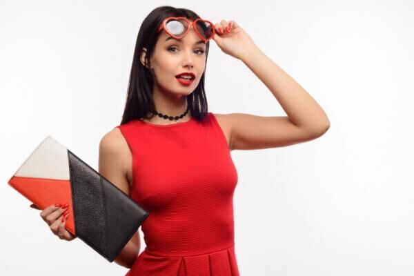 fashionable surprised young lady in a red dress and clutch holds on to sunglasses in the shape of a heart